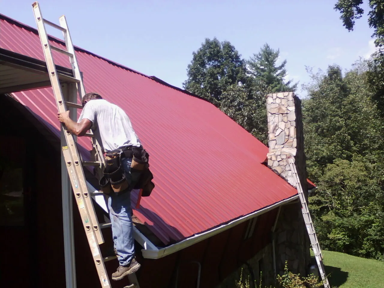 Roofer on a ladder beside a red metal roof and stone chimney — Soffit & Fascia Repair by Roofing Brothers in Anamosa